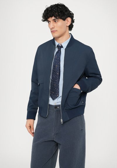 Námornícka bomber bunda so zipsom vpredu, rebrovanými manžetami, bočnými vreckami a hladkou textúrou, kombinovaná s svetlomodrou košeľou a vzorovanou kravatou.