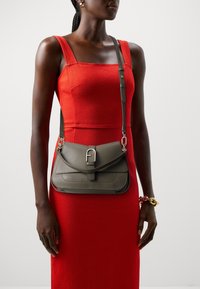 Olivinvihreä nahkainen olkalaukku, jossa on läppäkiinnitys, hopeiset yksityiskohdat ja säädettävä hihna, käytetty kirkkaan punaisen istuvan mekon päällä.