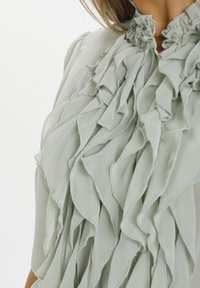 Blouse vert clair à volants présentant un tissu texturé, un col haut avec un bord froncé et de délicats volants verticaux sur le devant.