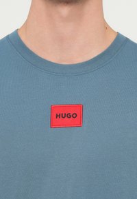 Kék pamut póló kerek nyakkivágással, a mellrészen egy piros téglalap alakú folttal, amelyen fekete betűkkel "HUGO" felirat található.