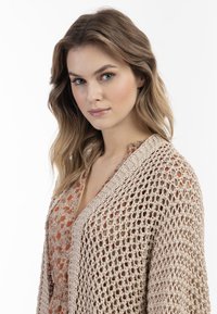 Beige gebreide cardigan met een open weefsel, gedragen over een bloemenpatroon jurk. Heeft een V-hals en getextureerde ribbels aan de randen.