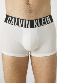 Hvite bomullsboksebriefs med en svart elastisk linning som har "CALVIN KLEIN"-logoen i hvitt. Myk tekstur, tettsittende design.