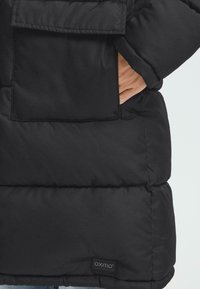 Sort pufferjakke med kviltet design, stor frontlomme og et logo-mærke på kanten. Materialet ser glat og isoleret ud.