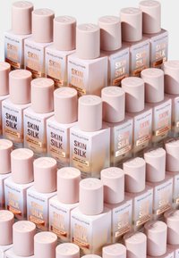 Staplade flaskor med Revolution Skin Silk. Flaskorna är rosa med rundade toppar, har klara etiketter och guldtext. Flera variationer syns.