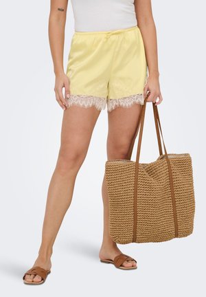 Kvinde iført gule shorts med blonder, hvid top, brune slippersandaler, holder en stor flettet taske ved siden af sig.