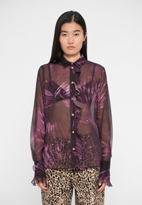 Blusa trasparente viola con stampa di foglie di palma, fronte arricciato e bottoni dorati; abbinata a pantaloni con stampa leopardata, dotati di polsini a campana.
