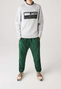 Osoba ubrana w jasnoszary sweatshirt Lacoste, ciemnozielone spodnie dresowe i beżowe sneakersy z wielokolorowymi detalami, stoi z rękami w kieszeniach.
