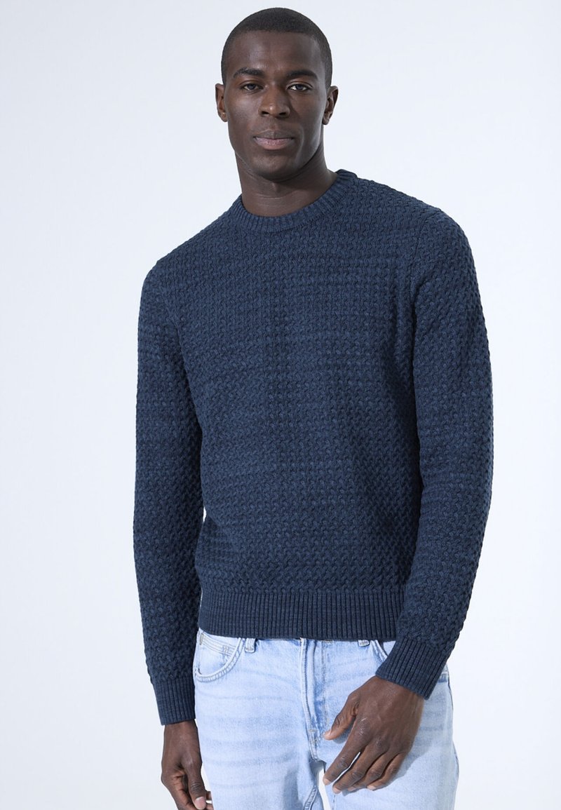 Marineblauwe gebreide trui met een textuurpatroon, ronde hals en geribde manchetten en zoom. Gecombineerd met lichtblauwe jeans.