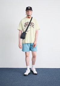  Lichtgele oversized t-shirt met gedrukte tekst, blauwe shorts, zwarte schoudertas, wit en groene sneakers, zwarte pet, binnen op een blauwe tapijt.