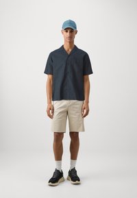 Cămașă navy cu mânecă scurtă, pantaloni scurți bej, adidași negri cu accente crem, asortați cu o șapcă de baseball bleu deschis.
