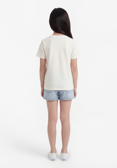 Enfant debout de dos, portant un t-shirt blanc uni à manches courtes, un short en jean bleu clair et des baskets blanches sur fond blanc.