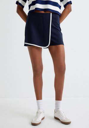 Jupe-culotte marine avec des bordures blanches et un design enveloppant, fabriquée en tissu doux. Portée avec une chemise rayée et des baskets blanches.