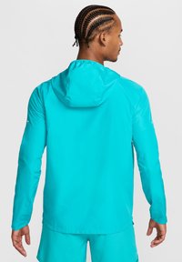 Lichtgewicht jas met capuchon in helder turkoois. Voorzien van lange mouwen, een gladde textuur en een verlengd achterpand. Geschikt voor actieve draagmomenten.