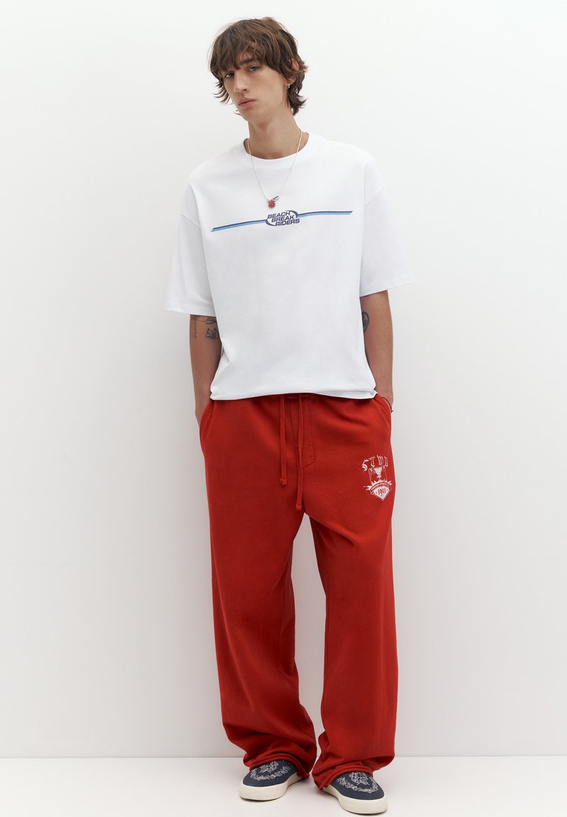 Jeune homme debout contre un mur blanc uni, portant un t-shirt graphique blanc, un pantalon rouge ample, des chaussures slip-on bleu marine à motifs et un collier.
