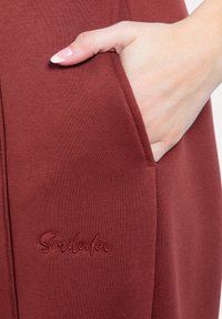 Bordový oděv s kapsou; má hladkou tkaninu a jemný vyšívaný detail "Smilestore" v odpovídající nitě.