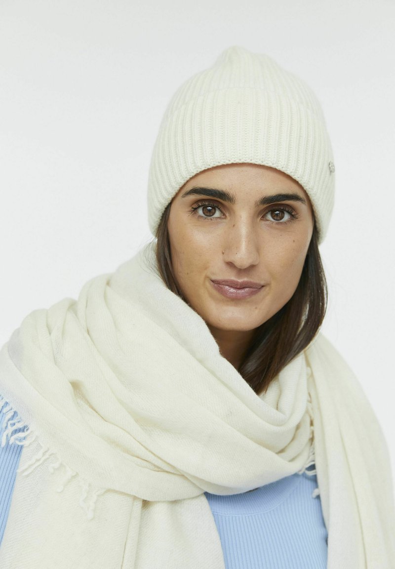 Weißer gerippter Strickbeanie mit spitzem Oberteil, kombiniert mit einem cremefarbigen, strukturierten Schal, der um den Hals drapiert ist. Darunter ein blauer strukturierter Pullover.