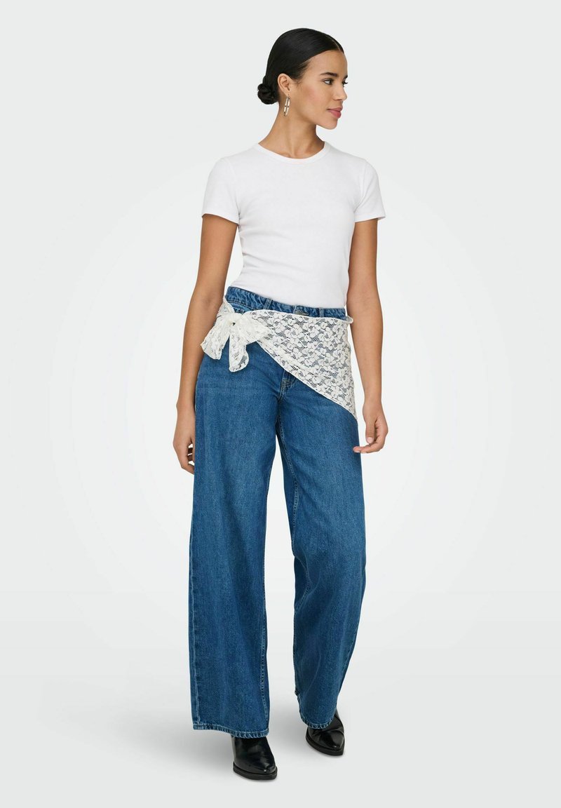Femme portant une chemise blanche à manches courtes, un jean bleu à jambes larges avec une ceinture en dentelle nouée à la taille, et des chaussures noires, debout et regardant vers la droite.