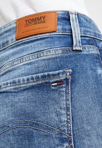 Acercamiento al bolsillo trasero de unos vaqueros de mezclilla azul con una etiqueta de cuero de Tommy Jeans y una pequeña etiqueta de bandera roja, blanca y negra en el bolsillo.