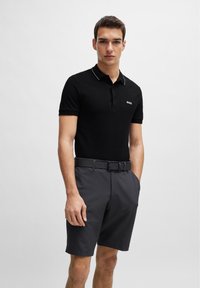 Svart polo-skjorte med hvit kragekant, med en logo på brystet. Kombinert med grå shorts og et matchende belte. Glatt stofftekstur.