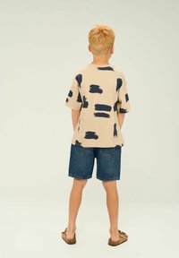 Camisa beige de manga corta con un patrón de pinceladas en azul marino, combinada con pantalones cortos de mezclilla azules. Los pantalones cortos son de longitud hasta la rodilla y tienen bolsillos traseros.
