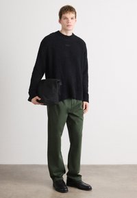 Pulover texturat negru, croială lejeră, asortat cu pantaloni verzi. Modelul ține o geantă neagră în mână și poartă pantofi negri. Fundal neutru.