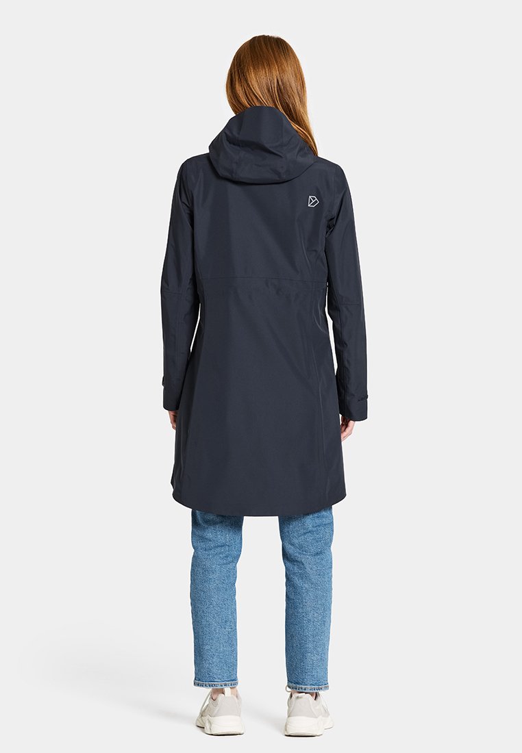 Marineblauer wasserdichter Mantel mit Kapuze, langen Ärmeln und tailliertem Schnitt. Verfügt über ein Logo auf dem Rücken und endet mittig am Oberschenkel. Kombiniert mit heller Jeans.