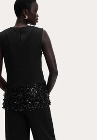 Top negro sin mangas con cintura ceñida, con un embellecimiento de lentejuelas negras texturizadas en el dobladillo y una pequeña abertura en la espalda.