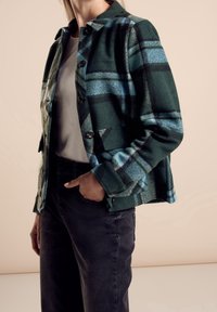 Zielono-niebieska kurtka w kratę z flaneli, z zapięciem na guziki, kołnierzem i dwiema przednimi kieszeniami. W połączeniu z czarnymi dżinsami.