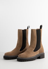 Bruine suède enkelboots met elastische zijpanelen, rubberen zool en contrasterende witte stiknaad. Stevig ontwerp met aantrektabs aan de bovenkant.