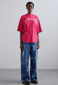 Červené oversized tričko s logom "Palm Angels", kombinované s modrými širokými džínsami so vzorom a čiernymi plochými topánkami. Hladké textúry, pohodlný štýl.