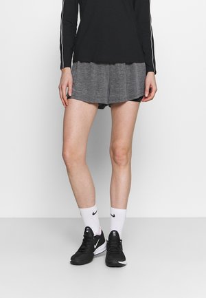 Person iført sort langærmet top, grå løse shorts, hvide Nike mellemlange sokker og sorte Nike Air sneakers, stående mod en ensartet hvid baggrund.