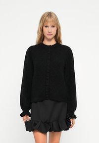 Svart stickad cardigan med ett diamantsmönster, rund halsringning och knäppning fram; matchad med en svart volangkjol. Mjuk textur, lös passform.