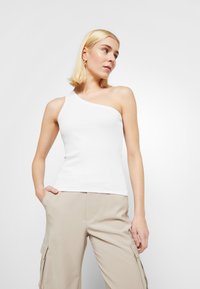 White, fitted tank top with one shoulder strap; smooth texture and minimalist design. Paired with beige cargo pants featuring pockets.