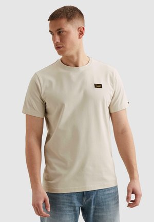 Joven con camiseta beige de manga corta con un pequeño parche de logo negro y amarillo, combinada con jeans azules, mirando hacia su izquierda.