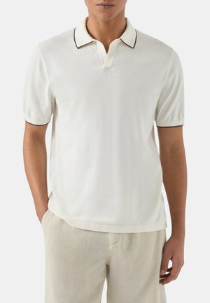 Uomo che indossa una polo bianca a maniche corte con rifiniture scure su collo e maniche, abbinata a pantaloni beige chiaro.