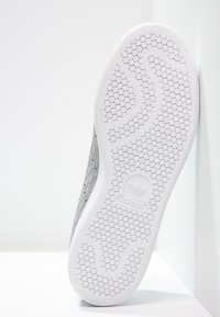 La suela de una zapatilla de goma blanca presenta un patrón de panal texturizado con un logotipo estampado en el centro, mostrada en un ángulo sobre un fondo claro.