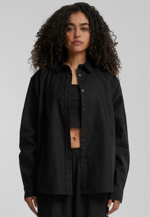 Zwarte button-up shirt van een gladde stof. Heeft een klassieke kraag, lange mouwen en een ontspannen pasvorm, gecombineerd met een zwarte tanktop eronder.