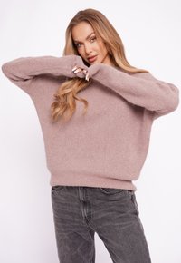 Jasnoróżowy, oversize'owy sweter wykonany z miękkiej, teksturowanej tkaniny z długimi, szerokimi rękawami. Połączony z ciemnoszarymi dżinsami, na białym tle.