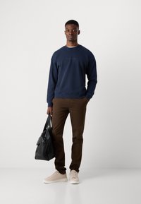 Navy sweatshirt with crew neckline, brown chinos, and light beige sneakers. Holding a black leather bag. Simple, casual outfit.