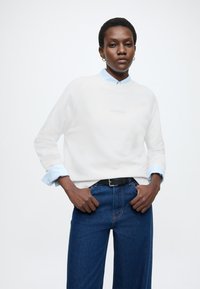 Fehér pulóver kerek nyakkivágással, világoskék galléros ing fölött viselve. Kék farmernadrág széles szárakkal és fekete övvel.