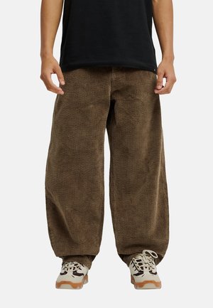 Persoon draagt wijde bruine corduroy broek en witte sneakers met lichtbruine zolen, staat met ontspannen armen langs het lichaam.