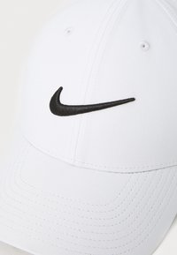 Hvid baseballcap lavet af tekstureret stof med et sort broderet Nike-logo på forsiden og syede detaljer på skyggen.