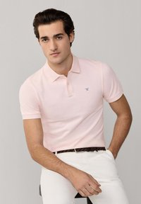 Homme portant un polo rose clair et un pantalon blanc, assis sur un tabouret devant un fond uni.