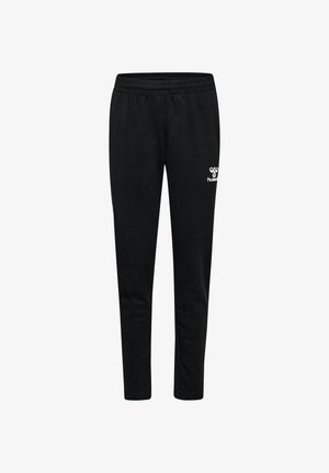 Mädchen trägt schwarze Sportbekleidung und Turnschuhe, steht breitbeinig mit einem leichten Lächeln vor einem schlichten hellen Hintergrund.