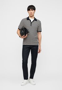 Mladić stoji, nosi polo majicu s crno-bijelim prugama kratkih rukava, tamne traperice, bijele tenisice, drži crnu kožnu torbu.