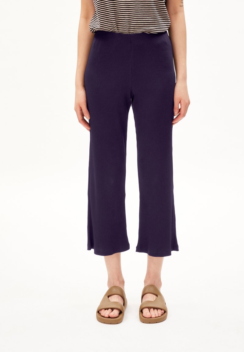 Personne portant un pantalon court large bleu marine, des sandales beige à enfiler et un haut rayé noir et blanc, debout devant un fond blanc.