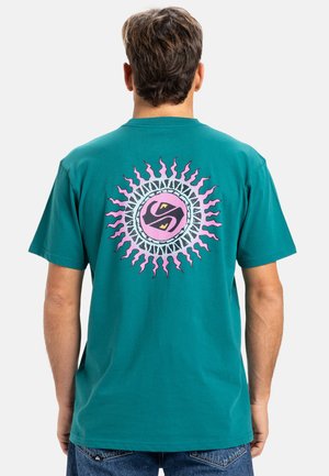 Teal katoenen t-shirt met een grote cirkelvormige afbeelding op de achterkant, met roze zonnestralen en een centraal logo in zwart en gele accenten.