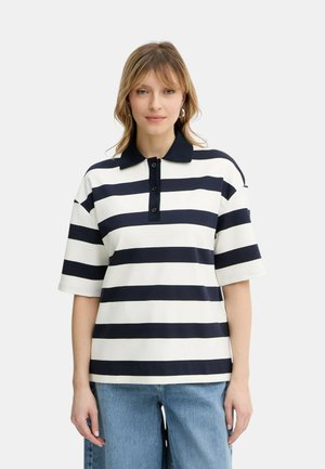 Donna che indossa una polo a maniche corte con strisce orizzontali bianche e nere e jeans blu, in piedi contro uno sfondo chiaro uniforme.