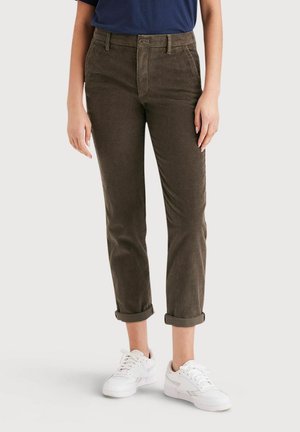 Personne portant un pantalon en velours côtelé marron avec les revers remontés, associé à des baskets blanches et un haut bleu marine, debout sur un fond uni.