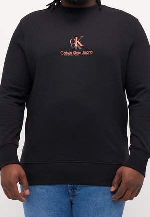 Mężczyzna w czarnej bluzie z okrągłym dekoltem z pomarańczowym logo Calvin Klein Jeans, w połączeniu z niebieskimi dżinsami, zwrócony przodem na jednolitym tle.